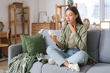 Young Asian woman with thyroid gland and pill bottle sitting on sofa at home