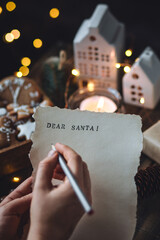 Christmas letter on craft paper to Santa Claus with text: Dear Santa. Cozy home interior with candles, lights, handmade toys, gingerbread cookies. Wishlist for presents from a child or adult