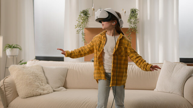 Funny happy kid girl playing online video 3d game using virtual reality headset child schoolgirl play in vr glasses at home in living room explore cyberspace world metaverse innovative technology AR