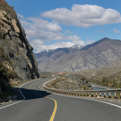 Long curved asphalt road running along the mountain pass. Crash barrier are along the road. Golden autumn roadscape with yellow trees and wild nature. Hilly taiga landscape in the distance