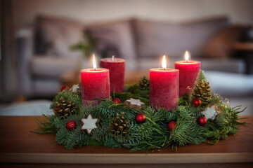 Third Advent, green wreath with Christmas baubles, cinnamon star cookies and red candles, three are lit, on a table in the living room, copy space, selected focus