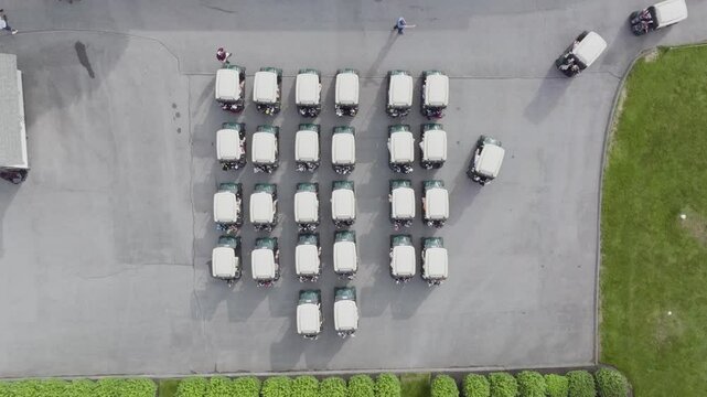 Birds-eye view of people getting into golf carts that sit in rows and slowly driving away from group in different directions. 