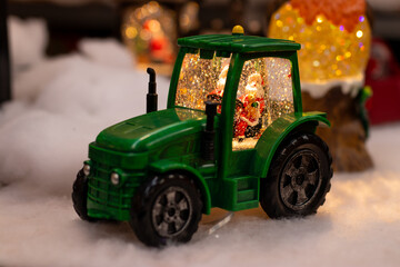 A festive green tractor ornament with Santa inside, surrounded by snow-like decor. The tractor glows warmly, creating a whimsical holiday ambiance with sparkling lights and snowy elements.