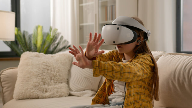 Funny caucasian kid girl child schoolgirl having fun in virtual reality headset at home couch playing online video game in world metaverse using innovative technology VR glasses exploring cyberspace