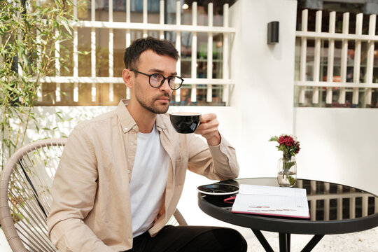 A young man in a stylish outfit sips coffee while seated outdoors at a cafe. A notebook on the table suggests a relaxing yet productive moment. Sunlight and plants add to the ambiance.
