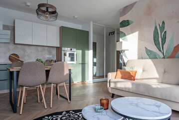 Modern living room with comfortable beige sofa and design wallpaper in green leaves on the wall. Stylish room with kitchen, table and chairs. Luxurious apartment.