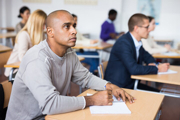 Focused adult latin american man listening to lecture in classroom with group. Postgraduate...