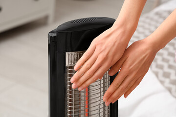 Young woman warming her hands near electric heater in bedroom, closeup