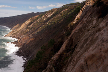 Dramatic coastal cliffs with rugged terrain, lush vegetation, and waves crashing against the rocky shore under a bright blue sky.