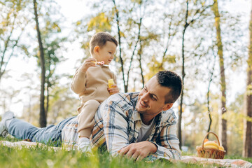 Fototapeta premium Happy attractive father and little son together sitting on blanket handsome boy blowing soap bubbles
