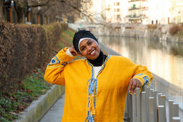 Black woman in yellow ethnic boho style clothing laughing joyfully and happily while standing by river in city park.