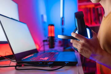 Close-up of a gaming laptop with RGB lighting on the keyboard, surrounded by colorful LEDs. A gamer holding a headset prepares for an online gaming session in a vibrant setup. © Marcio