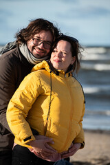 A couple stands embraced on a sandy beach on a chilly autumn day, the serene ocean stretching behind them as they share a quiet moment of love and warmth amidst the cool breeze and peaceful