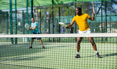 Sporty african american man padel tennis player trains on the court with a partner using a racket to hit the ball