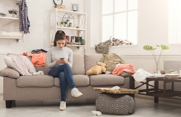 Obraz premium Desperate helpless woman sitting on sofa in messy living room. and chatting on mobile, surrounded by many stack of clothes. Disorder and mess at home, copy space