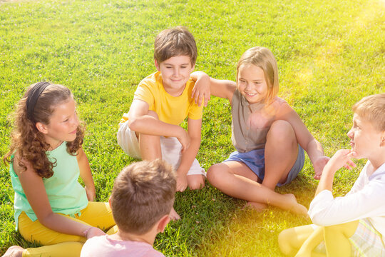 Cheerful preteen boys and girls spending time together outdoors on summer day, sitting on green lawn and talking ..