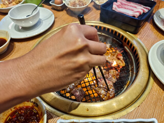 A close-up of a hand grilling meat slices on a barbecue grill using chopsticks, surrounded by side dishes and condiments on a wooden table, capturing a lively dining experience