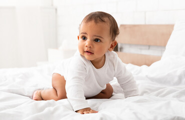 Fototapeta premium Cute African Little Baby Boy Crawling On Bed Looking Aside Posing In Bedroom At Home. Full Length Portrait Shot Of Adorable Toddler Kid. Sweet Childhood Moments, Child Care Concept