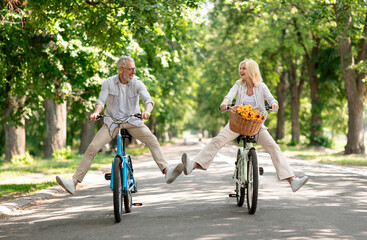 Active Retirement. Happy senior couple having fun while riding bicycles at summer park, cheerful mature man and woman cycling on retro bikes and laughing, enjoying outdoor leisure, copy space