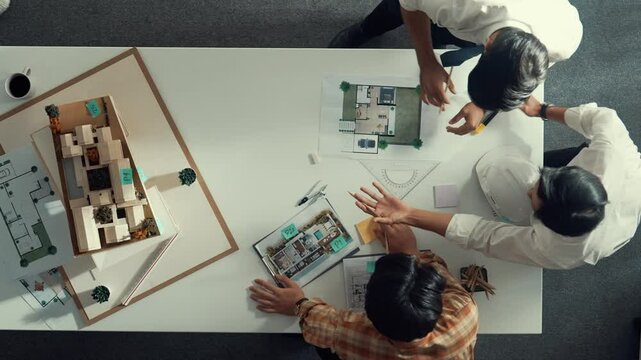 Group of skilled designer looking at model while discussing about building. Top view of architect engineer explain idea while talking and writing blue print scattering at meeting table. Alimentation.
