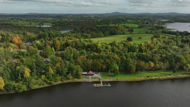 CAB&Uuml; by the Lakes, Killykeen Forest Park, County Cavan, Ireland, October 2023. Drone ascends tilting down over dock along lake with dense forested trees surrounding wood cabins and buildings.