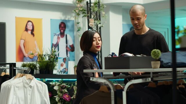 Asian store associate helps a young man client with style advice, embodying expertise and passion and dedication for the fashion industry. Woman improves customer satisfaction. Camera A.