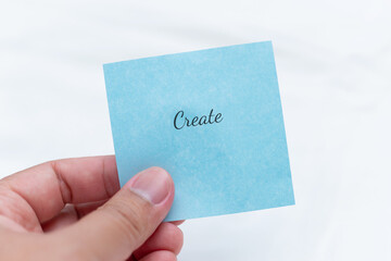 Hand holding blue note with text 'create' inspirational message minimalist setting close-up view...