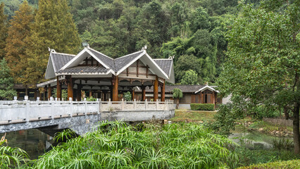 Gazebo with a triangular tiled roof in a park on the lake shore. A bridge with stone railings and...