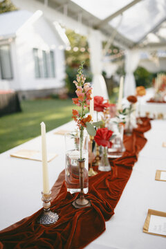 beautifully designed outdoor wedding reception featuring terracotta velvet accents, candlelit gold candlesticks, and floral arrangements with vibrant roses and snapdragons