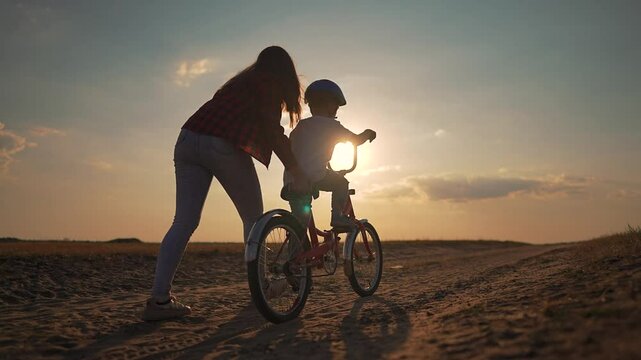 Mother helps son learning to ride bicycle at sunset. mother supports her son during his first bicycle lesson. Emotional family bonding moment as son is learning to ride bicycle scenic outdoor setting.
