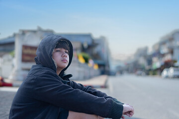 Asian young boy wearing a dark hooded jacket, sitting near a stone fence or barrier thoughtfully...