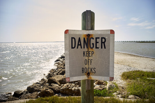 A warning sign stands on the edge of a rocky shoreline.