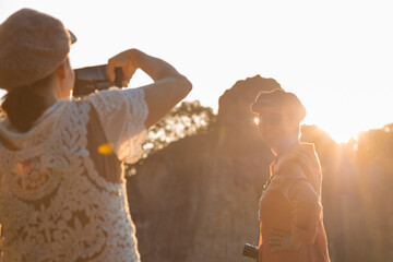 Two elderly women enjoying peaceful moment together outdoors during sunset, one taking a photo with smartphone and posing near serene water body, enjoying travel and capturing memories.