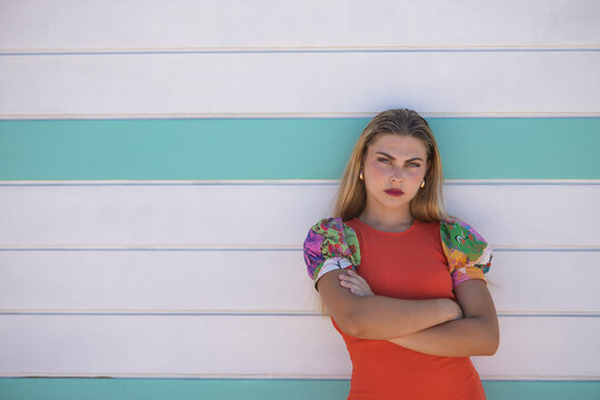 Young woman, beautiful, blue eyes, blonde, with orange colorful dress, arms crossed, empowered and independent leaning on a beach hut in the background. Concept beauty, fashion, trend, femininity. - Powered by Adobe