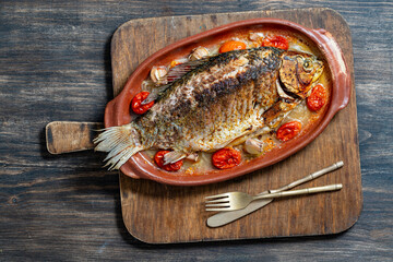 Delicious baked big fish crucian carp with spices in a clay plate and fried vegetables, closeup, top view