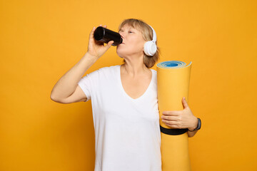 A woman drinks from a black cup while standing confidently with a rolled-up fitness mat under her...