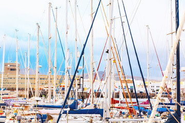 Densely packed marina with numerous sailboats docked closely together. Masts of the sailboats create a complex and visually striking pattern against the backdrop of buildings and a cloudy sky