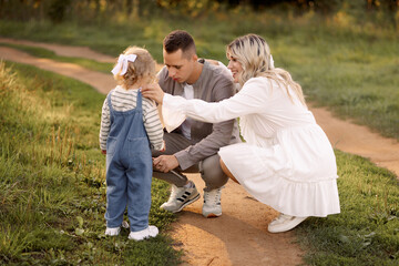 Fototapeta premium A young family with a toddler (around 2 years old) enjoys a moment in nature, smiling and blowing on dandelions. Perfect for concepts of family love, bonding, and childhood memories.