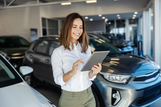Portrait of professional cheerful car saleswoman scrolling on tablet at car showroom while smiling at it.