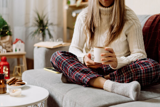 Cozy woman legs in knitted winter warm socks, sweater and checkered plaid drinking hot cocoa or coffee in mug, during resting on couch at home. Christmas holidays with candle, decor and cookies