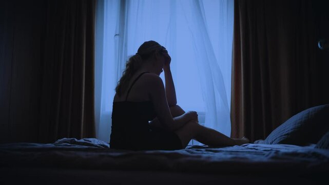 black silhouette upset unhappy woman sitting on bed dark room, cry wipes away tears with hand mood depression sadness emotion. Sad lonely Girl feels bad after party. dusk window curtains white fabric