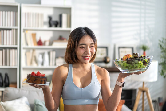 Young asian woman in sportswear making healthy lifestyle choice holding  salad bowl and slice of cake, fitness and health concept, balancing diet and exercise, home workout healthy living, motivation - Powered by Adobe