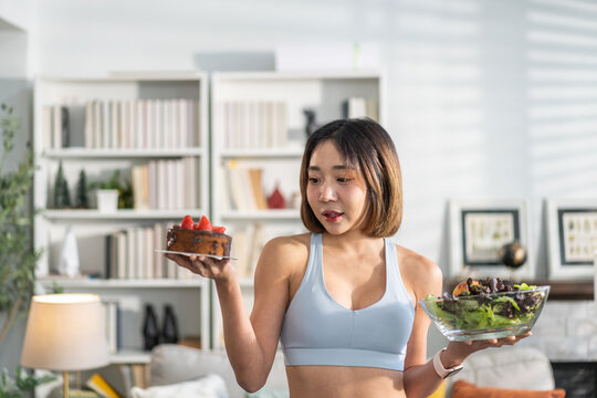 Young asian woman in sportswear making healthy lifestyle choice holding  salad bowl and slice of cake, fitness and health concept, balancing diet and exercise, home workout healthy living, motivation