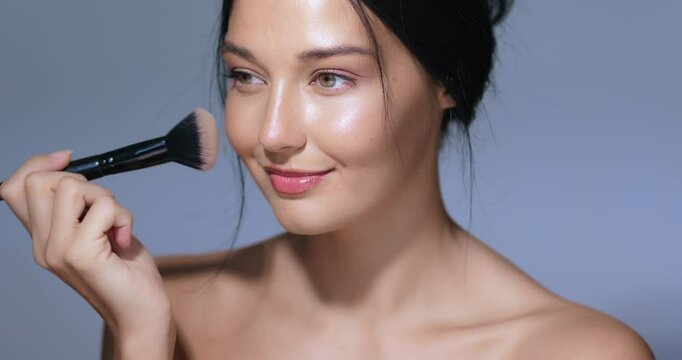 Beautiful woman with glowing skin and natural makeup applying blush with a makeup brush, smiling in a studio setting. Perfect for beauty tutorials, skincare ads, and cosmetics promotions. 