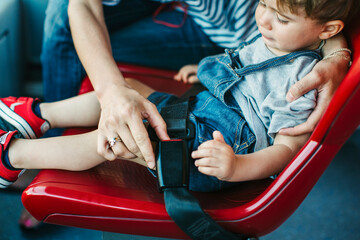 Mother fastens a seat belt for her son sitting on a bright red chair on a bus. The child looks focused and comfortable, ready for the journey ahead.