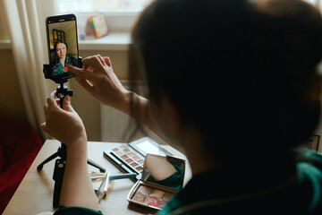 Plus size woman applying makeup on face with brush while recording vlog on smartphone for social...