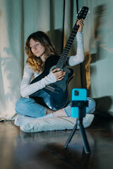 Young woman musician is recording a music video or online lesson with her acoustic guitar using her smartphone on a tripod