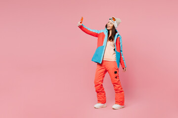 Full body skier young woman wear warm blue padded jacket ski goggles mask spend weekend winter season in mountains do selfie shot on mobile cell phone isolated on plain pink background. Hobby concept. © ViDi Studio