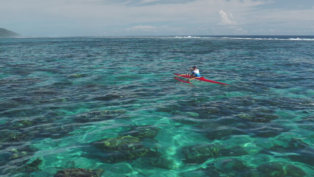Aerial drone captures a sportsman kayaking in the crystal-clear turquoise waters of Tahiti. French Polynesia. Showcasing the vibrant coral reef below and the lush green island in the distance