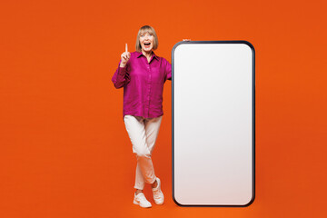 Full body elderly blonde woman 50s years old she wear purple shirt casual clothes big huge blank screen area mobile cell phone smartphone point index finger up isolated on plain red orange background.